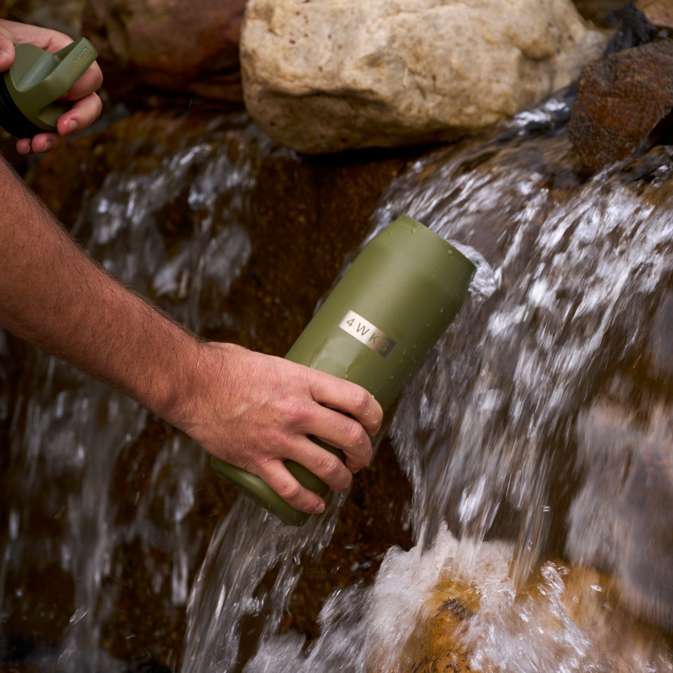 Person holding a green water bottle by a stream with rocks.